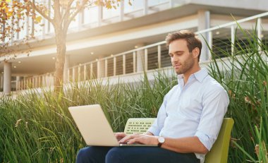 Genç işadamı modern bir ofis kompleksinin önündeki bankta oturuyor ve bir dizüstü bilgisayarla internette çalışıyor.