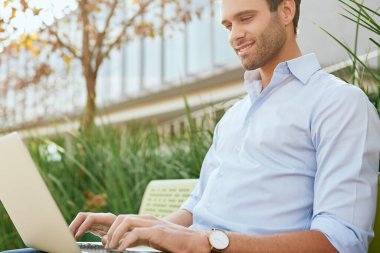 Ofisin dışındaki bankta oturan ve dizüstü bilgisayarla çalışan gülümseyen genç bir iş adamı.