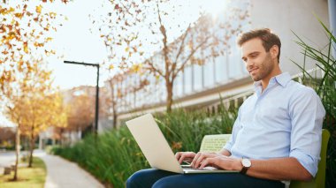 Modern bir ofis kompleksinin dışındaki bankta otururken bilgisayarla çalışan gülümseyen iş adamı  