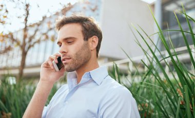 Genç işadamı ofis binasının dışında oturuyor ve cep telefonuyla konuşuyor.