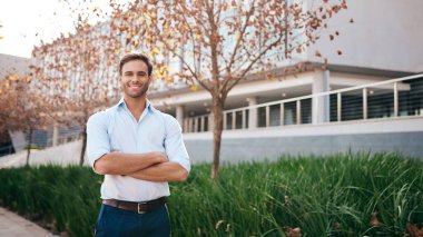 Bir ofis binasının önünde kollarını kavuşturmuş gülümseyen genç bir işadamının portresi.