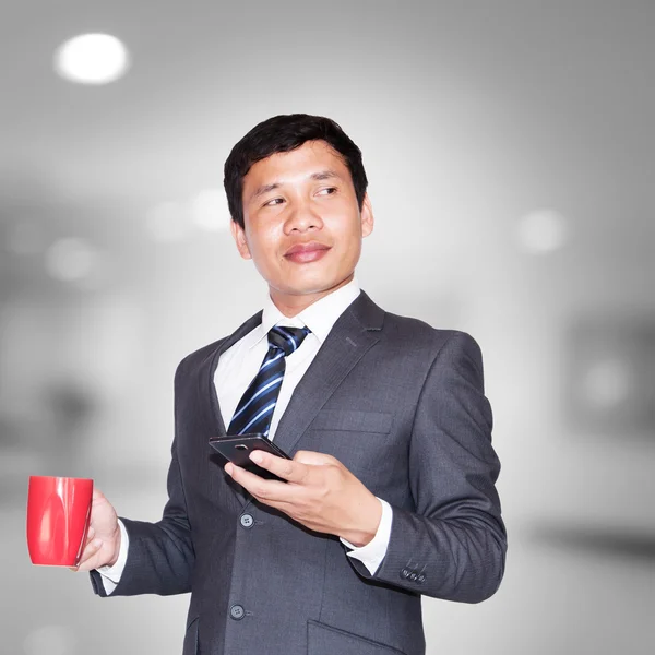 Hombre de negocios asiático con taza de café y el uso de teléfono inteligente mirando hacia otro lado en el espacio en blanco —  Fotos de Stock