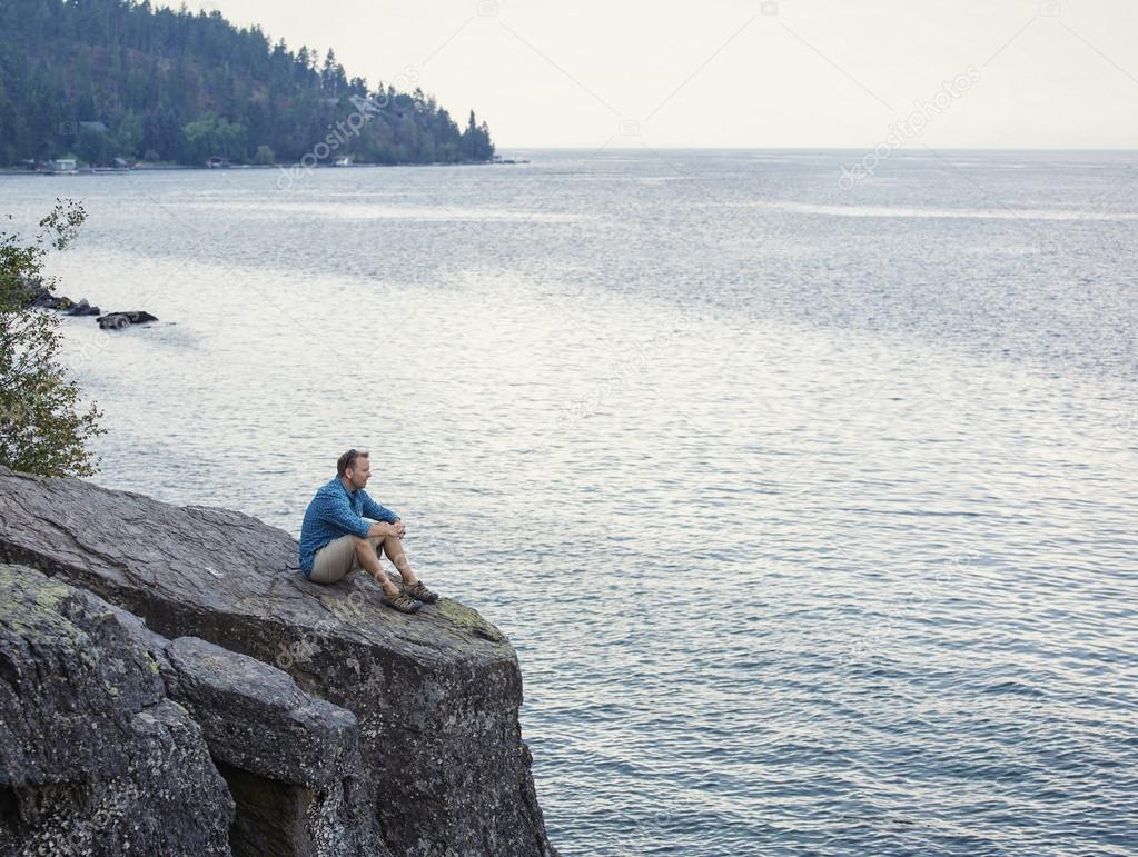 Man meditating and praying while sitting on the edge of large rock cliff overlooking the ocean.