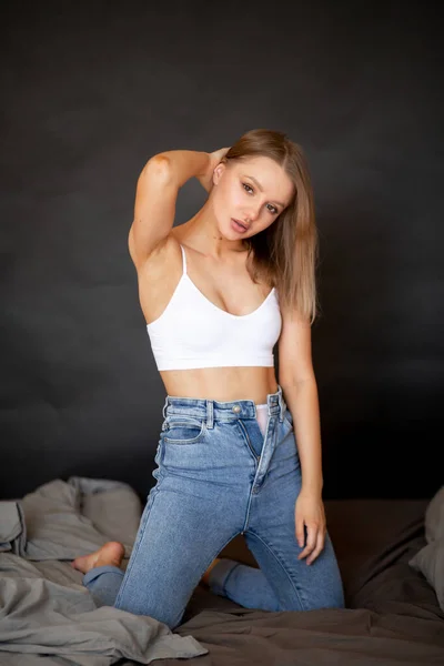 Mooi meisje in jeans en top zit op het bed en kijkt naar de camera — Stockfoto