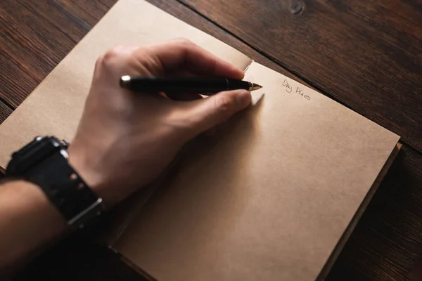 Een man schrijft in een notitieboekje Dagplannen met een pen op een donkere houten tafel. — Stockfoto