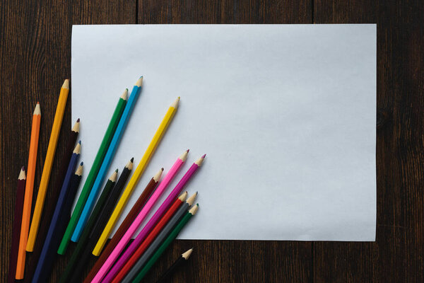 Чистый белый лист бумаги с цветными карандашами на темном деревянном фоне