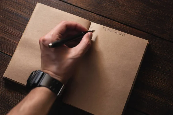 Een man schrijft in een notitieboekje Mijn plannen voor 2021 met een pen op een donkere houten tafel. — Stockfoto
