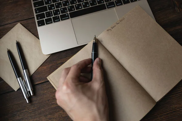 Een man schrijft in een notitieboekje Mijn plannen voor 2021 met een pen op een donkere houten tafel. — Stockfoto