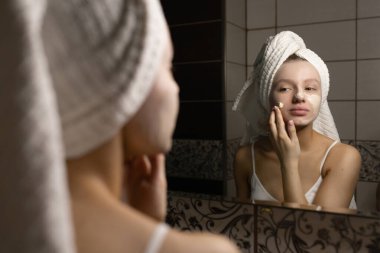 Banyoda kafasında havlu olan güzel beyaz bir kadın duştan sonra yüzüne krem sürüyor ve gülümsüyor. Kişisel bakım kavramı.