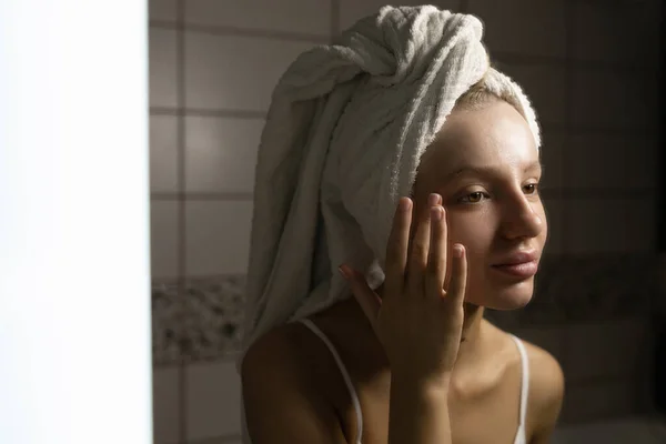 Όμορφη καυκάσια γυναίκα με μια πετσέτα στο κεφάλι της στο μπάνιο κοιτάζει στον καθρέφτη και απλώνει κρέμα στο πρόσωπό της. Φροντίδα του δέρματος προσώπου. — Φωτογραφία Αρχείου