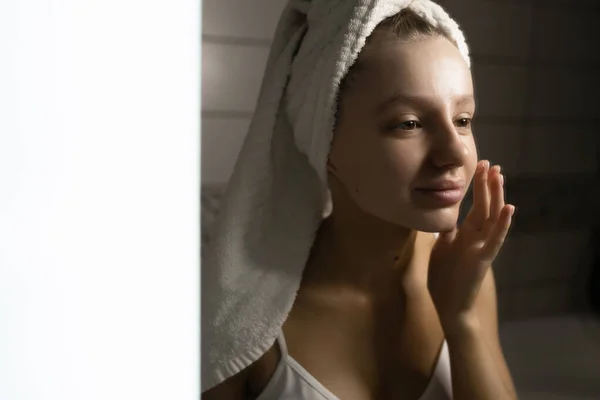 Hermosa mujer caucásica con una toalla en la cabeza en el baño aplica crema después de una ducha en la cara. Concepto de cuidado personal. Conservación de la belleza natural. — Foto de Stock