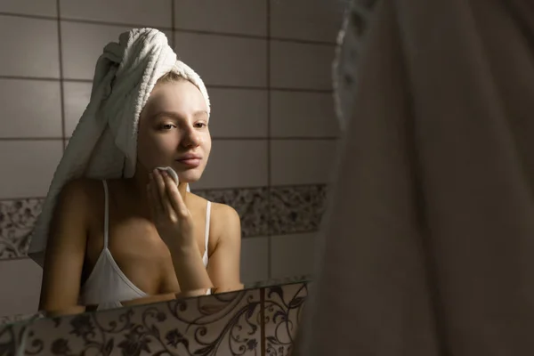 Hermosa mujer caucásica con una toalla en la cabeza en el baño después de una ducha se ve en el espejo y limpia la piel con un hisopo de algodón. Concepto de cuidado facial de la piel. — Foto de Stock