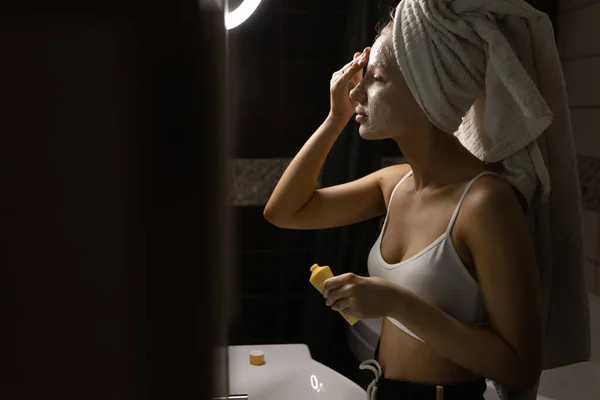 Une belle femme caucasienne avec une serviette sur la tête dans la salle de bain applique un masque crème sur son visage après une douche. Concept de soins de la peau. — Photo