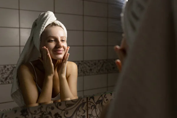 Όμορφη λευκή γυναίκα με μια πετσέτα στο κεφάλι της στο μπάνιο αφού ένα ντους κοιταχτεί στον καθρέφτη, χαϊδεύει το πρόσωπό της με τα χέρια και χαμογελά. Έννοια φροντίδας δέρματος. — Φωτογραφία Αρχείου