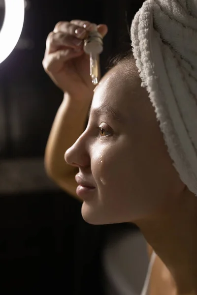 Hermosa mujer caucásica con una toalla en la cabeza en el baño después de una ducha se aplica suero en la cara con una pipeta. Cuidado facial de la piel. — Foto de Stock