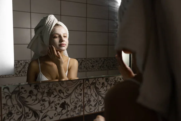 A beautiful Caucasian woman with a towel on her head in the bathroom applies a cream mask to her face after a shower, looks in the mirror and smiles. Facial skin care concept. — Stock Photo, Image