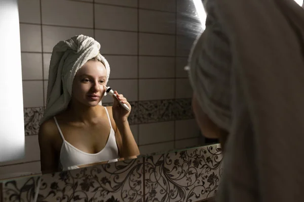 Belle femme caucasienne avec une serviette sur la tête dans la salle de bain applique de la crème sous les paupières. Concept de soins personnels. Préservation de la beauté naturelle. — Photo