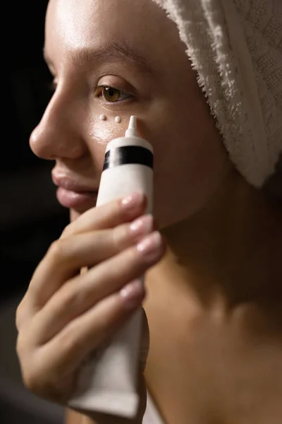 Hermosa mujer caucásica con una toalla en la cabeza en el baño aplica crema debajo de los párpados. Un plan cerrado. Concepto de cuidado de la piel. Conservación de la belleza natural. — Foto de Stock