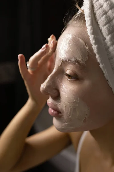 A beautiful Caucasian woman with a towel on her head in the bathroom applies a cream mask to her face after a shower. Close plan. Skin care concept. — Stock Photo, Image