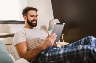 Sabahları yatakta yatarken dijital tablet kullanan pijamalı yakışıklı bir genç.
