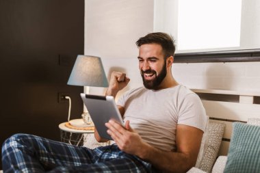 Sabahları yatakta yatarken dijital tablet kullanan pijamalı yakışıklı bir genç.