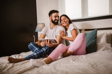 Evde yatakta dijital tablet kullanan gülümseyen rahat genç çift.