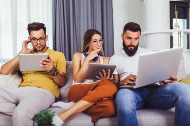 Arkadaşlar evdeki oturma odasındaki koltukta otururken dijital cihazlarını kullanıyorlar.