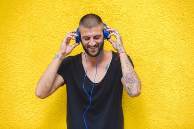 Dövmeli adam sarı bir duvarda kulaklık kullanarak müzik dinliyor.