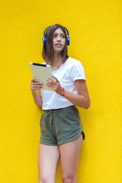 Young woman is listening to music on digital tablet using headphones on a yellow wall