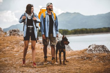 Sırt çantalı yürüyüşçüler ve dobermanları dağ gölünün keyfini çıkarıyorlar.