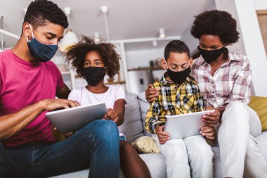 Evdeki aile. Afrikalı Amerikan ailesi koruyucu maskeler takıyor. Evlerinde dijital tablet kullanıyorlar..