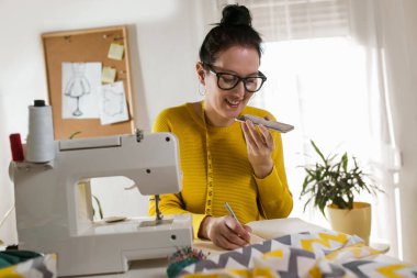 Evinde dikiş makinesine telefon kullanarak dikiş diken bir kadın..