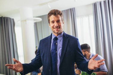 İş adamı lideri, arka planda iş adamları çalışırken modern ofiste kameraya bakıyor. Takım çalışması konsepti.