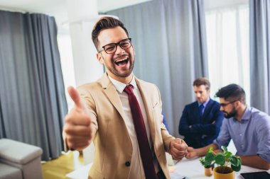 İş adamı lideri, arka planda iş adamları çalışırken modern ofiste kameraya bakıyor. Takım çalışması konsepti.