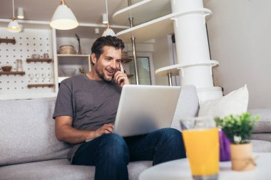 Oturma odasındaki kanepede oturmuş dizüstü bilgisayar kullanan, gündelik giysiler giyen çekici bir gencin portresi.