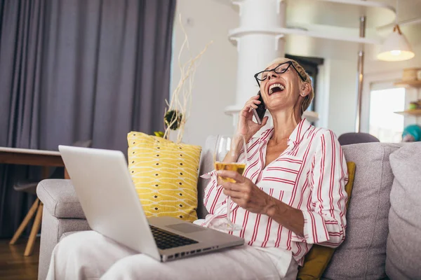 Happy middle aged woman with laptop computer, sitting at the couch while talking to someone on phone.