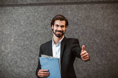 Genç, mutlu iş adamı elinde bir pano ile modern binanın önünde başparmağını kaldırıyor.