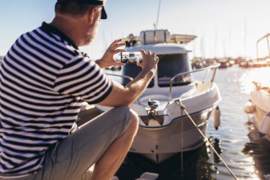 Denizci gömleği ve şapkası giymiş, akıllı telefon kullanarak denizin kenarında duran olgun bir adam.