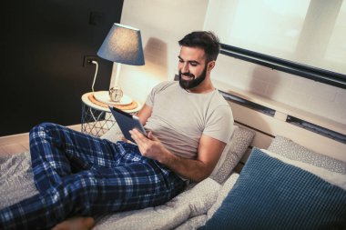 Elinde tabletle yatakta yatan gülümseyen bir erkek. Ekrana zevkle ve neşeyle bakıyor.