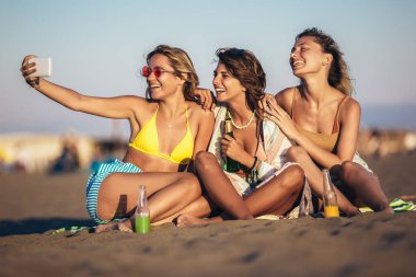 Kumsalda oturup bira ve meyve suyu içen mutlu genç kadınlar telefonla selfie çekiyorlar. Bir grup arkadaş plaj tatilinde eğleniyor..