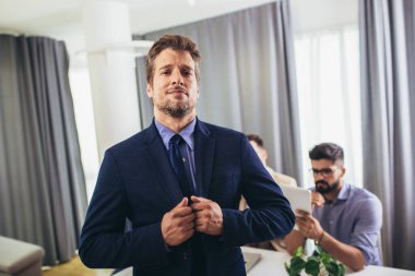 İş adamı lideri, arka planda iş adamları çalışırken modern ofiste kameraya bakıyor.. 