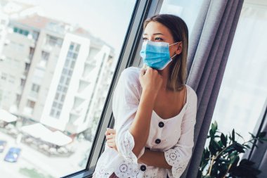 Tıbbi koruyucu maskeli genç bir kadın pencereden dışarı bakıyor.