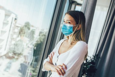 Tıbbi koruyucu maskeli genç bir kadın pencereden dışarı bakıyor.