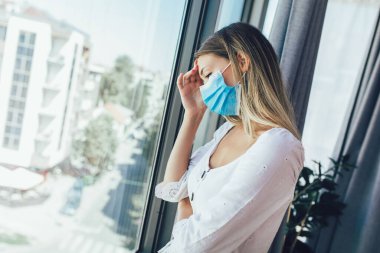 Tıbbi koruyucu maskeli genç bir kadın pencereden dışarı bakıyor.