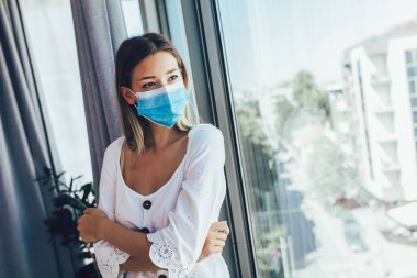Tıbbi koruyucu maskeli genç bir kadın pencereden dışarı bakıyor.