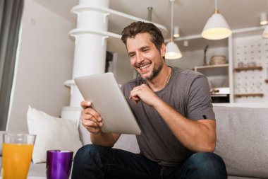 Yakışıklı adam dijital tablet kullanıyor ve evinde kanepede dinlenirken gülümsüyor.