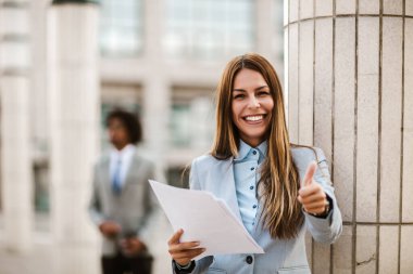 İş kadını portresi açık havada gülümsüyor iş belgelerini tutuyor, başparmağını göster.