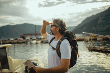 Gülümseyen turist olgun bir adam harita ve sırt çantasıyla denizin kenarında duruyor..