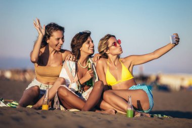 Kumsalda oturup bira ve meyve suyu içen mutlu genç kadınlar telefonla selfie çekiyorlar..