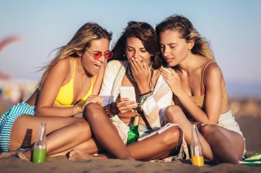 Kumsalda oturan mutlu genç kadınlar akıllı telefon kullanarak bira ve meyve suyu içerler.. 